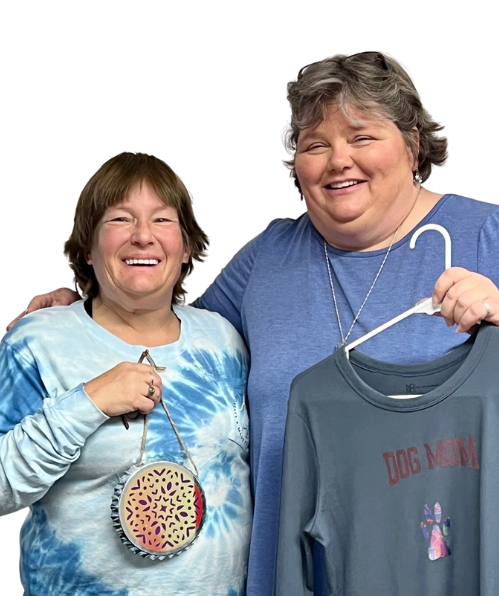 Two women stand together, arms around each other, holding craft items: a gray T-shirt that says 